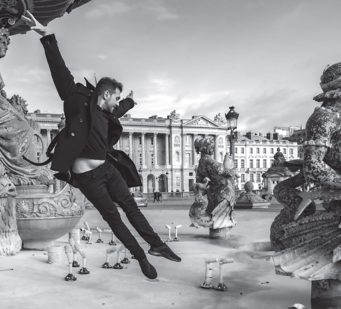 A man in a dark coat leaps mid-air between the statues of a fountain on a city square, black-and-white photograph